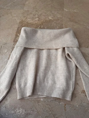 Beige off shoulder stickad tröja - Mysig beige stickad tröja med bred off shoulder-krage och långa ärmar. Tröjan har ribbstickade muddar vid ärmslut och nederkant samt en lös passform. Perfekt för lager på lager och en avslappnad stil.