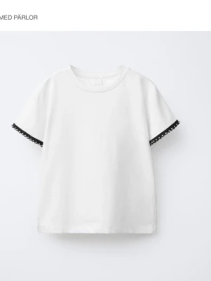 Zara T-shirt med detalj på ärm - En basic t shirt fast med söta pärldetaljer på ärmarna från Zara. Kan inte hitta den på hemsidan så den är troligtvis slutsåld. Köpte den för flera år sedan men har bara legat i garderoben så den är oanvänd. Storleksvis är den rekommenderad till barn, passar mig som är 18 och 160 cm och vanligtvis har xs-s i kläder. Den är en blandning av vit och beige i färgen.