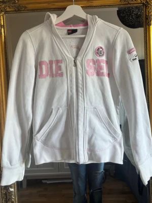 Vit hoodie med dragkedja från Diesel - Vit hoodie från Diesel med dragkedja framtill och rosa detaljer. Stor DIESEL-text i rosa på bröstet och broderat blommotiv samt text på ryggen. Två fickor fram och huva. Materialet är mjukt och skönt, troligen bomullsblandning. Snygga patches på ärmen och cool streetwear-vibe.