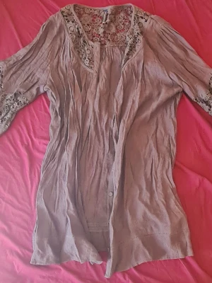 Beige blus med spetsdetaljer - Säljer en beige blus med vackra spetsdetaljer vid axlar och ärmslut. Blusen har knappar framtill och ett luftigt, löst sittande fall. Materialet känns lätt och skönt, perfekt för en avslappnad men ändå stilren look.