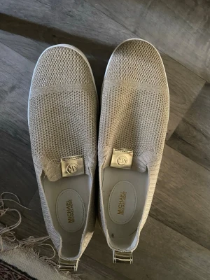 Beige slip-on sneakers från Michael Kors - Stilrena slip-on sneakers från Michael Kors i beige meshmaterial med diskret logodetalj framtill. Skorna har rund tå och platt sula, perfekt för en avslappnad och trendig look.