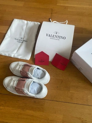 Valentino Garavani sneakers rosa/vit - Snygga sneakers från Valentino Garavani i vitt läder med bred rosa rem över vristen och nitar på sulan. Klassisk låg modell med vita skosnören och subtil logga på plösen. Perfekta för dig som vill ha en exklusiv touch till din outfit.