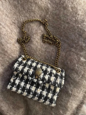 Kurt Geiger väska  - Säljer en svartvit rutig handväska i tweed med guldfärgad kedjerem och dekorativt snäckformat spänne framtill. Väskan har en klassisk, lite boxig form och materialet ger en lyxig vintagekänsla. Perfekt för dig som gillar statement-accessoarer.