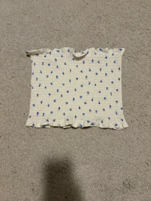 Blommig vit bandeau topp med volang - Supersöt bandeau topp i vitt med små blå blommor och volangkant upptill och nedtill. Smockad och stretchig för en bekväm passform. Perfekt för sommardagar och enkel att matcha med jeans eller kjol.