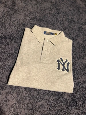 Grå piké från Polo Ralph Lauren NY - Snygg grå pikétröja från Polo Ralph Lauren med New York Yankees-logga på bröstet och klassisk krage med knappar. Tillverkad i mjuk bomull och har korta ärmar. Perfekt för dig som gillar sportig och clean stil. Kom gärna med frågor och funderingar pris ej hugget i sten.