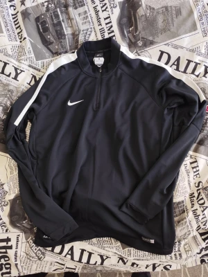 Svart Nike träningsjacka med dragkedja - Svart långärmad träningsjacka från Nike med vit logga på bröstet och vita ränder längs axlarna. Jackan har halv dragkedja framtill och är tillverkad i ett lätt, tekniskt material som andas. Perfekt för träning eller uppvärmning.