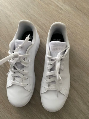 Vita Adidas sneakers i läder - Fräscha helvita sneakers från Adidas med klassisk låg profil. Skorna har ovandel i slätt läder, vita skosnören och diskret Adidas-logga på plösen och innersulan. Snygg minimalistisk design som passar till det mesta. Yttersulan är också vit för en clean look.