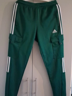 Gröna cargobyxor från Adidas - Snygga gröna cargobyxor från Adidas med vita ränder längs sidorna och praktiska fickor på benen. Byxorna har en sportig look och är tillverkade i ett mjukt, stretchigt material som ger skön passform. Perfekta för en avslappnad streetwear-stil.