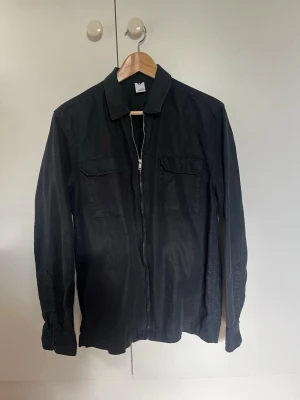 Svart overshirt med dragkedja - Svart overshirt med dragkedja framtill och två bröstfickor med lock. Skjortan har klassisk krage och långa ärmar med manschett. Perfekt att bära öppen eller stängd för en avslappnad stil.