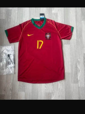 Portugal Nike fotbollströja Ronaldo 17 - Röd Portugal fotbollströja från Nike med gröna detaljer vid hals och ärmslut. Nummer 17 i gult framtill och Portugals landslagsemblem på bröstet. Klassisk V-ringning och korta ärmar, perfekt för fotbollsträning eller match.
