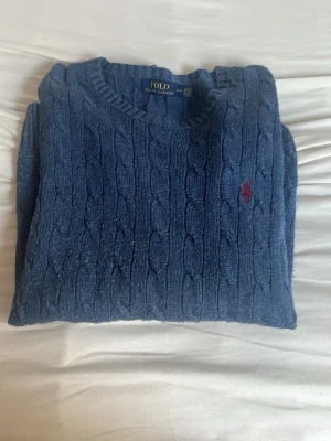 Blå kabelstickad tröja Polo Ralph Lauren - Kabelstickad blå tröja från Polo Ralph Lauren med rund halsringning och klassisk röd logga broderad på bröstet. Tröjan har en tidlös design och är perfekt för lager-på-lager. Materialet är mjukt och bekvämt.