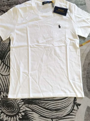 Vit t-shirt från Polo Ralph Lauren - Klassisk vit t-shirt från Polo Ralph Lauren med den ikoniska svarta logobrodyr på bröstet. T-shirten har rund hals och korta ärmar, tillverkad i mjuk bomull för en skön känsla. Perfekt basplagg med stilren design. Passar fint mellan 160-170 cm.