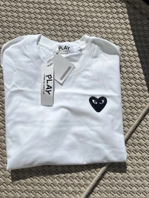 Vit Comme des Garçons PLAY t-shirt - Vit t-shirt från Comme des Garçons PLAY med klassisk svart hjärtdetalj på bröstet. T-shirten har rund halsringning och korta ärmar. Tillverkad i mjuk bomull för en skön och clean look.