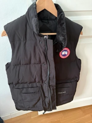 Svart dunväst från Canada Goose - Snygg svart dunväst från Canada Goose med klassisk logga på bröstet. Västen har hög krage, dragkedja och knappar framtill samt två stora fickor. Perfekt för lager-på-lager och håller dig varm under kyliga dagar.