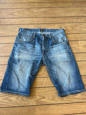 Blå jeansshorts från Lee - Säljer ett par klassiska blå jeansshorts från Lee med fem fickor och bälteshällor. Shortsen har en avslappnad passform och tydliga slitningar för en cool, casual look. Perfekta för sommaren och lätta att matcha med olika stilar.