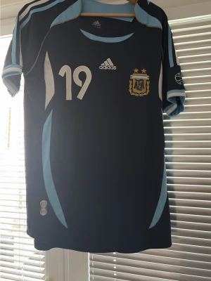 Argentina Messi matchtröja Adidas - Säljer en blå Argentina fotbollströja från Adidas med nummer 19 och Messi på ryggen. Tröjan har ljusblå och vita detaljer, officiellt landslagsmärke på bröstet och klassiska Adidas-ränder på axlarna. Perfekt för dig som älskar fotboll och Messi.