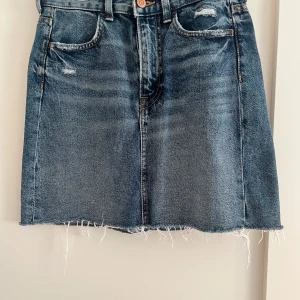Helt ny blå jeanskjol  - Blå jeanskjol i klassisk modell med fem fickor, slitna detaljer och rå, fransig kant nedtill. Kjolen har normal passform och stängs med knapp och dragkedja framtill. Perfekt till sommaren och enkel att styla med olika toppar. Helt ny blå jeanskjol från HM med högmidja. Storlek 34. Inga defekter 