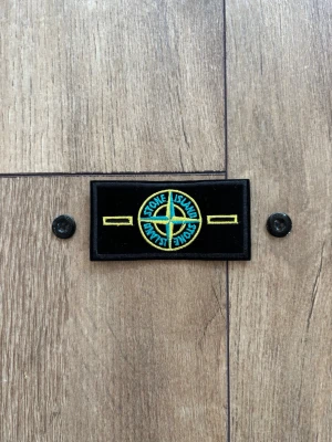 Stone Island Classic Badge / Patch – Helt ny & Äkta (Med knappar) - Säljer en helt ny och oanvänd klassisk Stone Island-badge med tillhörande originalknappar. Den har den ikoniska designen med gul och grön kompasslogga på svart botten. Perfekt om du har tappat bort din gamla patch eller vill ha en fräsch reservdel till din Stone Island-jacka, sweatshirt eller hoodie. I absolut nyskick! 