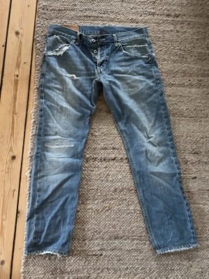 Blåa Dondup jeans  - Säljer ett par blå jeans med slitningar och råa kanter. Jeansen har knappgylf, fem fickor och coola detaljer som slitna partier och ljusare tvätt. Perfekta för en avslappnad streetstil.