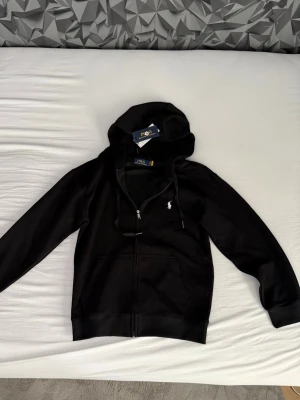 Svart hoodie från Polo Ralph Lauren - Snygg svart hoodie från Polo Ralph Lauren med dragkedja framtill och klassisk vit logga på bröstet. Hoodien har justerbar huva med snören och två fickor fram. Tillverkad i mjukt material som känns skönt mot huden. Helt ny med ettiket.