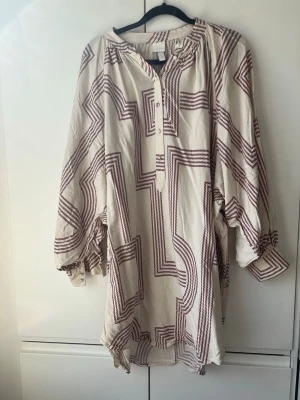 Vit och vinröd mönstrad klänning/tunika - Säljer en luftig vit klänning med vinröda grafiska ränder. Klänningen har lång ballongärm, v-ringad hals med knappar och avslappnad passform. Perfekt för dig som gillar unika mönster och bekväma plagg.