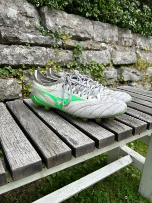 Gråa Mizuno fotbollsskor med neon - Säljer ett par gråa Mizuno fotbollsskor med snygga neongröna detaljer och klassisk snörning. Skorna har en låg profil och är tillverkade i syntetmaterial med mönstrad ovandel för bättre bollkontroll. Perfekta för dig som vill sticka ut på planen!