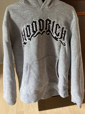 Grå HOODRICH hoodie med tryck - Snygg grå hoodie från HOODRICH med stor svartvit logga på bröstet och text på huvan. Klassisk känguruficka framtill och mjukt material som känns skönt mot huden. Perfekt för dig som gillar streetwear och vill ha en avslappnad look.