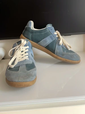 Maison Margiela blå sneakers - Maison Margiela sneakers i blå mocka och läder med vita snören och gummisula. Klassisk låg modell med stilren design och diskreta sömmar. Perfekta för dig som gillar en clean och trendig look. Priset e verkligen inte hugget i sten speciellt inte under ett snabbköp