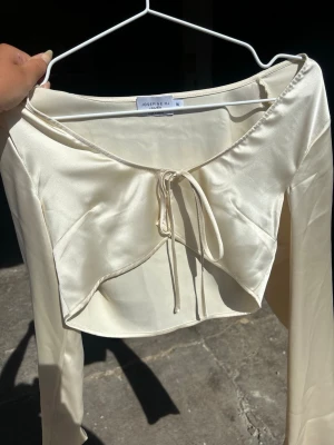 Champagnefärgad satin bolerotopp - Snygg champagnefärgad bolerotopp från Josefine HJ i glansigt satinliknande material. Modellen är croppad med långa ärmar och har knyt framtill för en trendig look. Perfekt att styla över ett linne eller klänning.