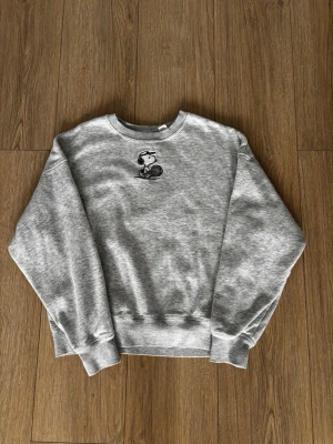 Grå sweatshirt med Snoopy-tryck - Mysig grå sweatshirt med rund hals och ett litet Snoopy-tryck på bröstet. Tröjan har lång ärm och ribbade muddar vid ärmslut och nederkant. Perfekt för en chill och avslappnad stil. Obs ! Storlek barn 13 år /158 cm 