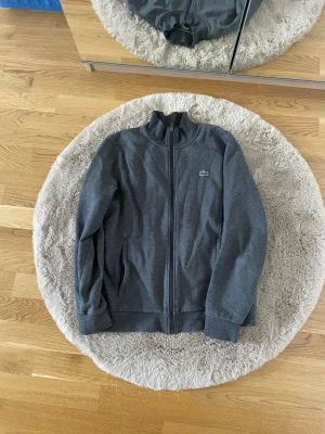 Grå zip-hoodie från Lacoste Sport - Mörkgrå hoodie från Lacoste Sport med dragkedja och ståkrage. Klassisk krokodillogga på bröstet och mjukt, vitt fleecefoder på insidan. Perfekt för en sportig och avslappnad stil. Tillverkad i bomullsblandning för extra komfort.