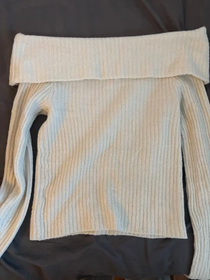Off shoulder stickad tröja cremevit - Trendig off shoulder stickad tröja i cremevit färg med ribbad struktur. Långa ärmar och bred krage som viks ner över axlarna. Perfekt för dig som vill ha en stilren och mysig look till hösten.