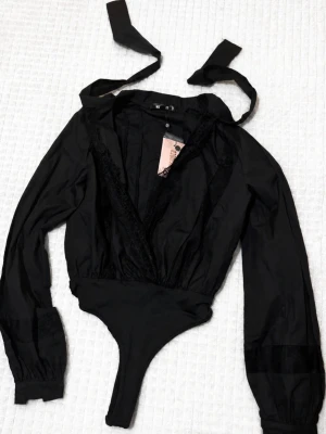 🖤 Snygg svart body från missguided - Nyskick!🖤 - Elegant svart body med djup v-ringning, fina spetsdetaljer och blusliknande överdel med långa ärmar. Modellen har även två dekorativa snören som knyts runt halsen för en feminin och elegant look✨️  Helt ny och oanvänd - lapp/tag sitter fortfarande kvar.  🖤Färg: Svart 🖤Skick: Ny med lapp kvar 🖤Modell: Body med djup v-ringning, spets och knytning i halsen 🖤Märke: missguided (köpt på nelly) 🖤Storlek: XS (kan passa s beroende på önskad passform)  💌 Skriv vid frågor eller fler bilder 