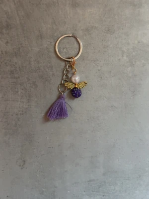 Nyckelring med lila tofs och ängel - Unik nyckelring med en lila tofs och en dekorativ ängelberlock i guld, pärla och lila glittrande pärla. Metallring i silverfärg. Perfekt accessoar för att piffa upp väskan eller nyckelknippan. Kan även hängas på träns eller grimma🤗 Har även massor av färger eftersom att jag gör dessa själv.