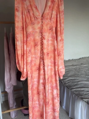 Populär adoore klänning storlek 32 - Säljer en lång klänning i ljusa orange och rosa toner med blommigt mönster. Klänningen har v-ringning, långa puffärmar och dekorativa knappar framtill. Perfekt för sommarens alla tillfällen! Använd 2 gånger, inga defekter och har med extraknapp samt prislapp. Storlek 32 men passar även mig som har både 32/34💕slutsåld 