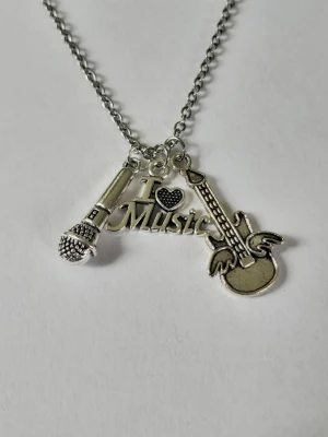Halsband med musiktema i silverfärg - Snyggt halsband i silverfärg med tre hängen: en mikrofon, en elgitarr med vingar och en textplatta med 'I ♥ Music'. Perfekt för dig som älskar musik och vill visa det med din stil. Kedjan är tunn och smidig, och hänget ger en cool och personlig touch till din outfit. Materialet rostfritt stål. 