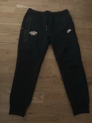 Svarta mjukisbyxor från Nike X syna world - Svarta mjukisbyxor från Nike med vita loggor på framsidan och dragsko i midjan. Byxorna har ribbade muddar vid bensluten och en extra ficka med dragkedja på ena benet. Perfekta för en avslappnad och sportig stil.