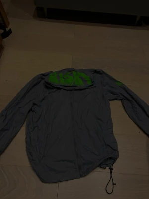 Risky vindjacka - Snygg grå vindjacka från Risky med dragkedja framtill och huva. Jackan har en grön Nike-logga på huvan och är tillverkad i lätt, vindtätt material. Justerbar nederkant med dragsko för bästa passform. Perfekt för dig som vill ha en sportig och stilren look.