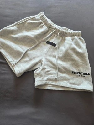 Grå Essentials Fear of God shorts - Snygga ljusgrå shorts från Essentials Fear of God med svart logga på benet. De har elastisk midja med snörning och är gjorda i mjuk bomull för maximal komfort. Perfekta för en avslappnad och trendig look.
