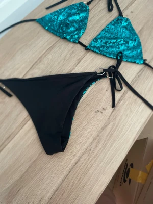 Turkos paljettbikini med knyt - Snygg bikini med turkosa paljetter på både överdel och underdel. Triangelformad topp med knyt i nacken och ryggen. Underdel med svarta sidor och knytband samt ringdetaljer. Perfekt för sommarens strandhäng!