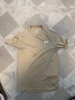 Beige tränings-tshirt från Under Armour - Snygg beige tränings-tshirt från Under Armour med diskret geometriskt mönster. Tillverkad i ett lätt och tekniskt material som andas, perfekt för gymmet eller löprundan. Klassisk rund hals och korta ärmar. Logga i vitt på bröstet.
