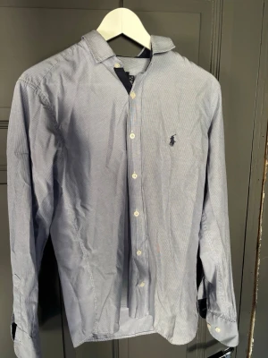 Blå randig skjorta från Ralph Lauren - Klassisk blå och vit randig skjorta från Ralph Lauren med lång ärm och knappar framtill. Skjortan har den ikoniska loggan broderad på bröstet och är tillverkad i mjuk bomull. Perfekt för en stilren och avslappnad look.