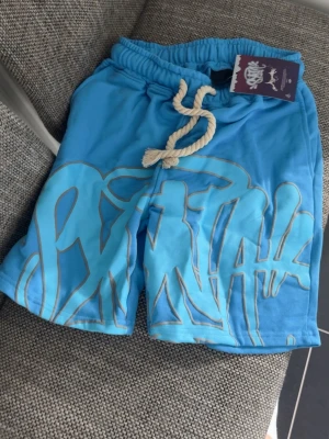 Syna world T-shirt och Shorts  - Snygga blå shorts med coolt graffiti-inspirerat tryck framtill och grov snörning i midjan. Shortsen har elastisk midja och en bakficka. Perfekta för sommaren och riktigt sköna att bära. Passar L/M (DEN ÄR EJ ANVÄND) behövs bara strykas, då den legat i garderoben hela tiden