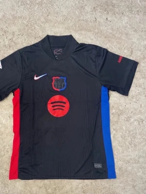 Svart FC Barcelona matchtröja Nike - Säljer en svart FC Barcelona matchtröja från Nike med klubbmärke och Spotify-logga på bröstet. Tröjan har röda och blå detaljer på sidorna, korta ärmar och UNHCR-tryck på ryggen. Perfekt för dig som älskar fotboll och vill ha en snygg supportertröja.