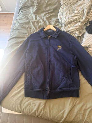 Marinblå jacka från Polo Ralph Lauren - Snygg marinblå jacka från Polo Ralph Lauren med dragkedja framtill och två fickor. Jackan har ribbade muddar vid ärmslut och nederkant samt en liten gul Polo-logga på bröstet. Perfekt för dig som gillar klassisk och stilren design.