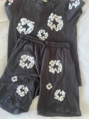 Denimtears - Säljer ett svart set med t-shirt och shorts, dekorerat med vita blommönster i olika storlekar. T-shirten har rund hals och korta ärmar, shortsen har resårmidja med snörning. Perfekt för en avslappnad och trendig look.