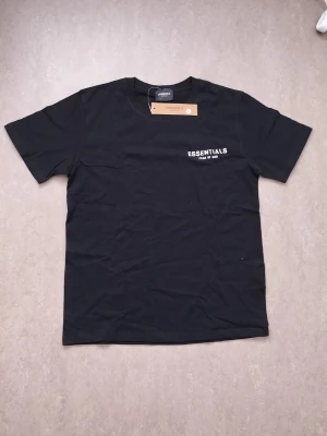 Svart Essentials t-shirt Fear of God - Snygg svart t-shirt från Essentials Fear of God med vit logga tryckt på bröstet. Klassisk passform med rund hals och korta ärmar. Tillverkad i mjuk bomull som känns skön mot huden. Perfekt för en clean och stilren look.