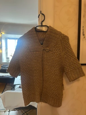 Beige stickad kofta med kort ärm - Beige stickad kofta med korta ärmar och stor krage. Koftan har ett grovt stickat mönster och stängs med en knapp framtill. Perfekt för lager-på-lager och chill stil. Materialet känns mjukt och mysigt.