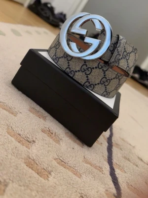 Gucci beige GG-bälte med silverdetalj - Säljer ett klassiskt Gucci-bälte i beige med det ikoniska GG-mönstret och stor rund silverfärgad logga som spänne. Bältet är gjort i skinn och har en lyxig känsla, perfekt för att lyfta vilken outfit som helst. 105cm