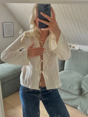 Vit blus med knytning framtill - Trendig vit blus med tre knytband framtill och lätt utsvängda ärmar. Blusen har en luftig känsla och är tillverkad i ett tunt, strukturerat tyg. Perfekt att styla med jeans för en avslappnad look.